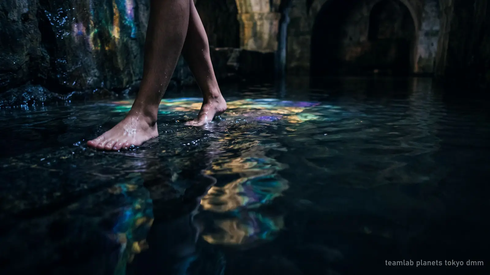 Illustration : des pieds nus avançant dans une eau jusqu'aux genoux, dans une salle sombre baignée de lumière colorée, le type d'espace aquatique qui a fait la renommée de teamLab Planets